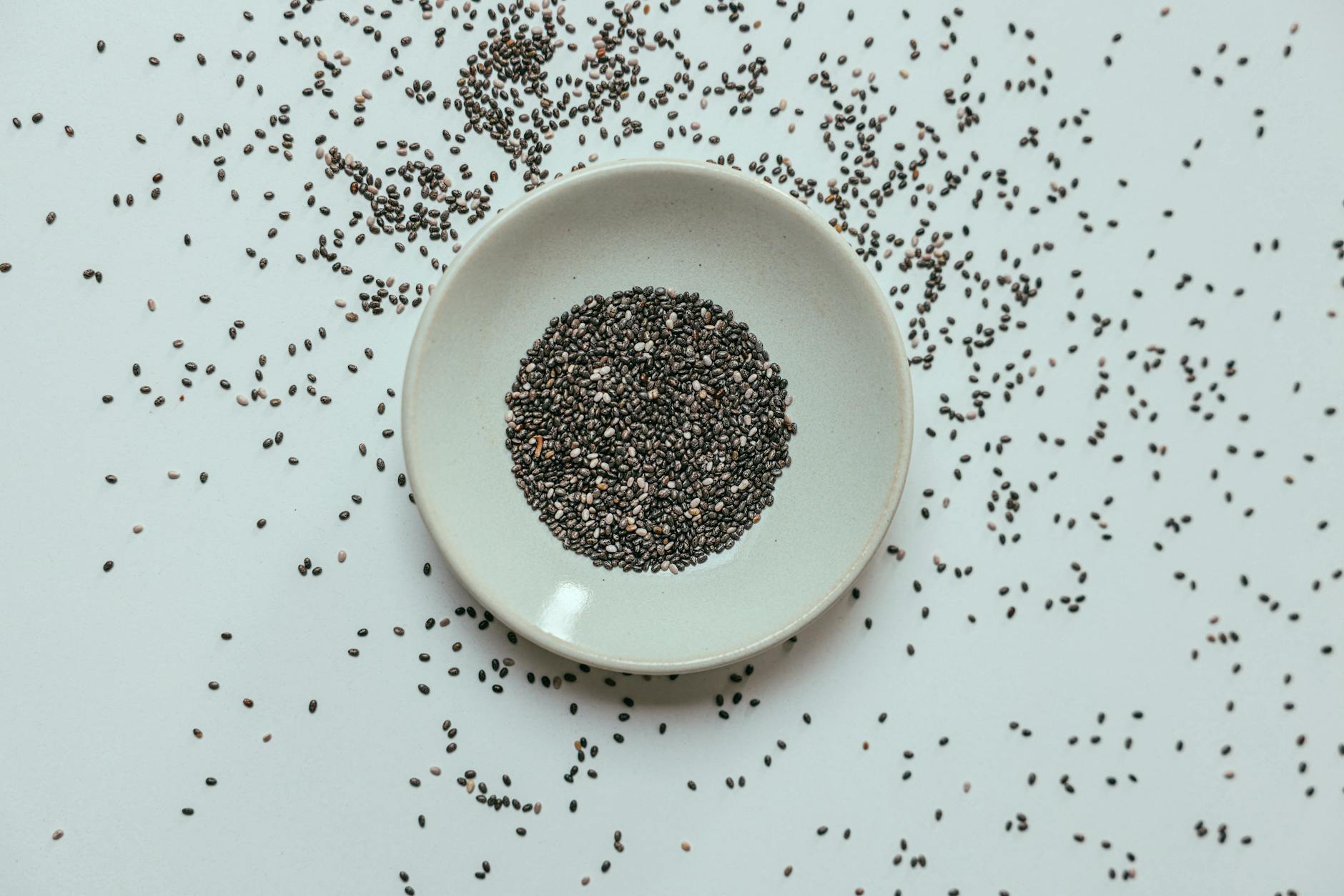 Top view of chia seeds in a white bowl with scattered seeds around on a flat surface.