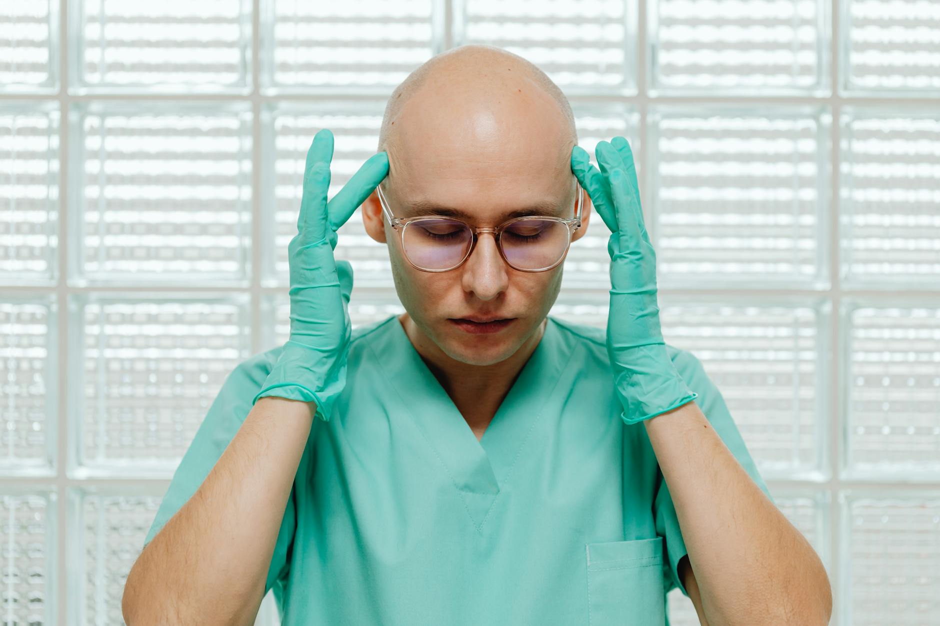 A surgeon in green scrubs appears stressed, holding their head with gloved hands indoors.