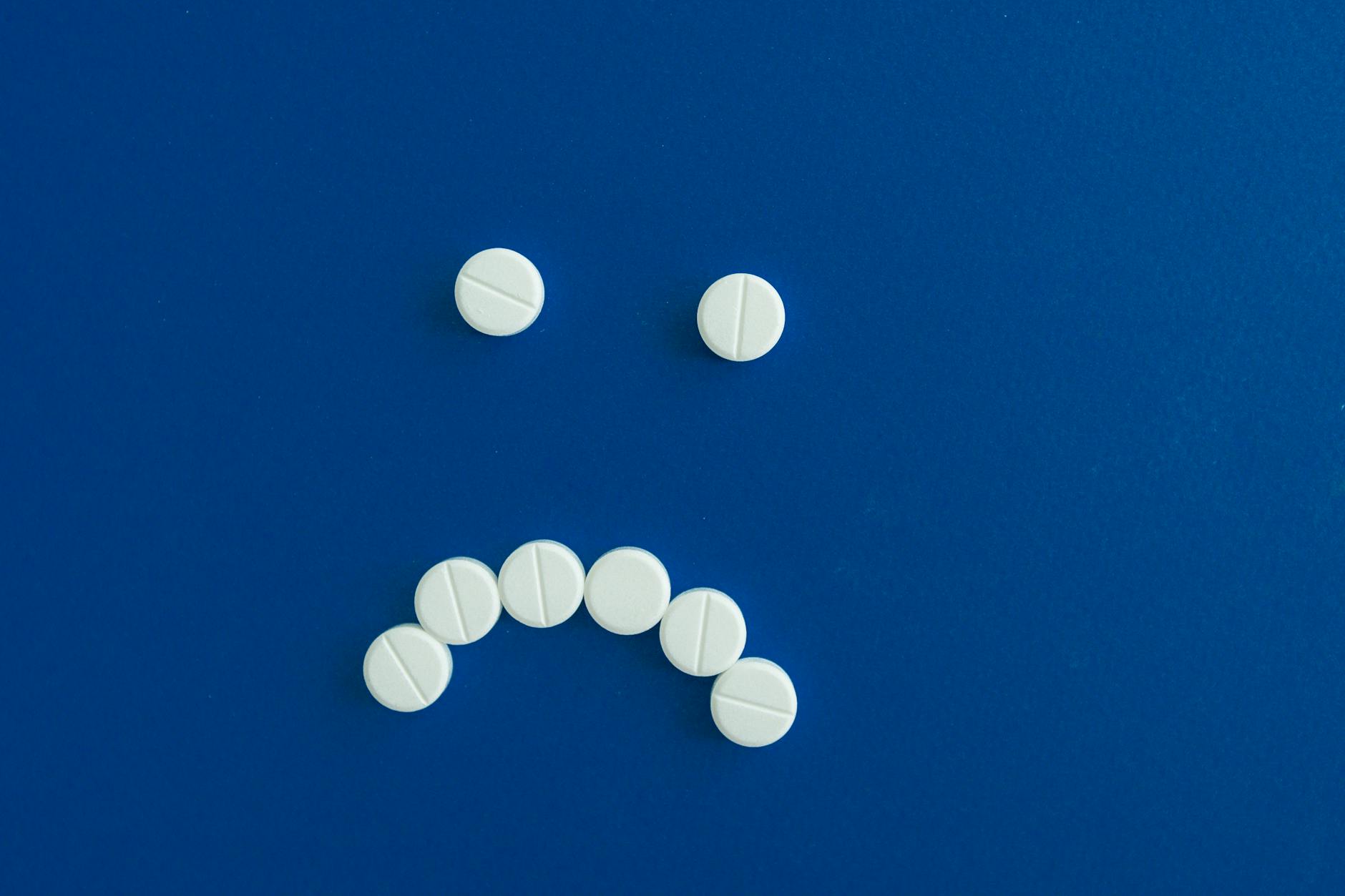 White pills arranged in a sad face shape on a solid blue background.