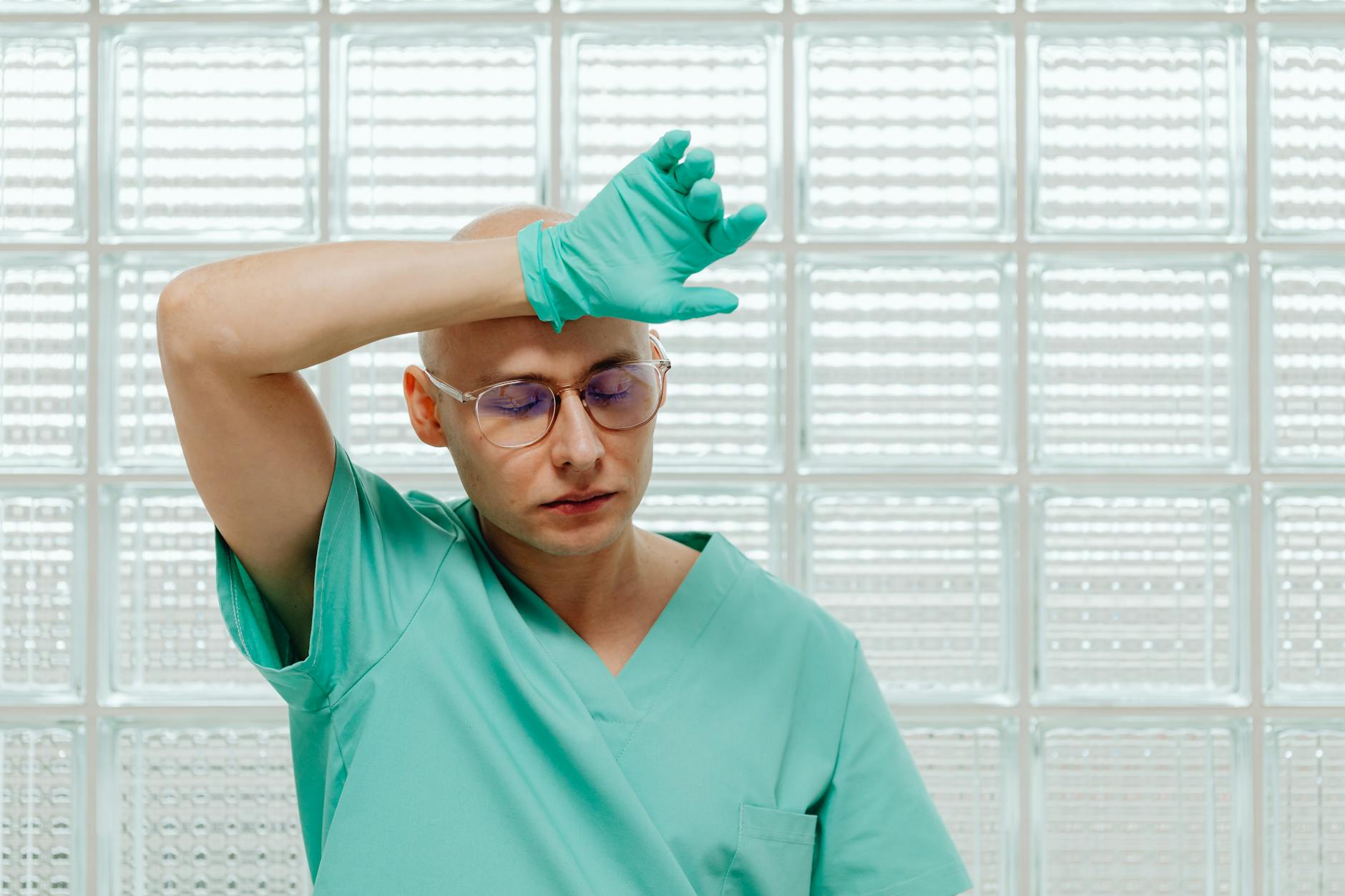 Tired male doctor wearing green scrubs and gloves, wiping brow in a medical setting.